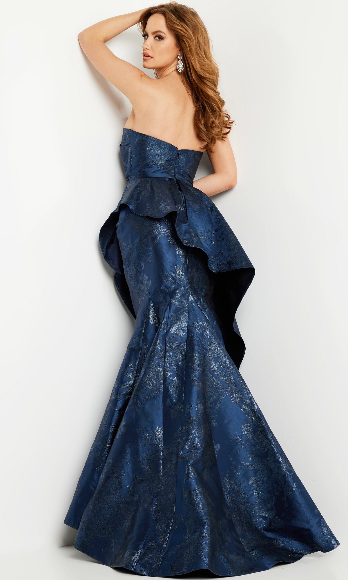 Woman wearing the Jovani 23624 Long Formal Dress, a strapless dark blue floor-length gown with peplum detail, posing with one arm raised and looking over her shoulder.
